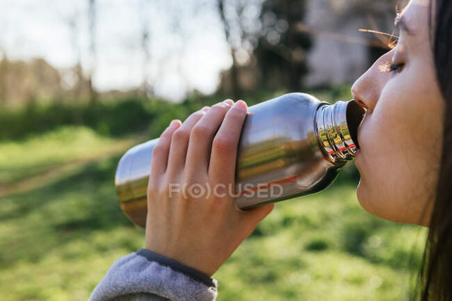 Seitenansicht einer Wanderin, die im Wald steht und beim Wandern frisches Wasser aus einer Metallflasche genießt — Stockfoto