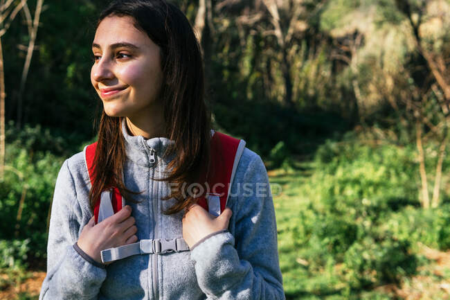 Щаслива оптимістична молода жінка ходить в активному одязі з рюкзаком, насолоджуючись подорожжю в зеленому лісі в сонячний день — стокове фото