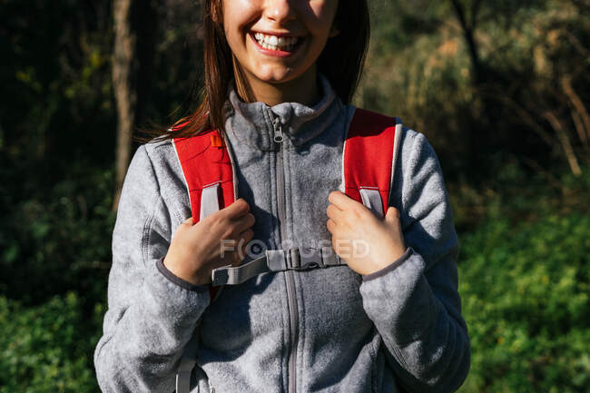 Anónimo feliz optimista joven excursionista en ropa deportiva con mochila disfrutando de un viaje en un bosque verde en un día soleado - foto de stock