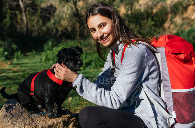 Seitenansicht der fröhlichen jungen Frau mit Rucksack streichelt niedlichen kleinen Hund beim Ausruhen beim Wandern im Wald — Stockfoto