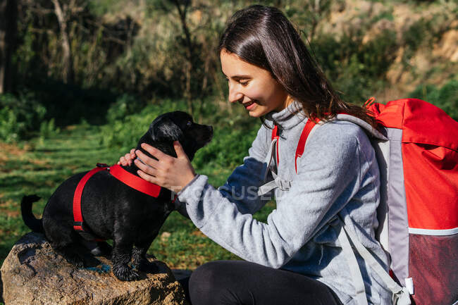 Vista laterale di allegra giovane donna con zaino accarezzando simpatico cagnolino mentre riposa durante le escursioni nella foresta — Foto stock