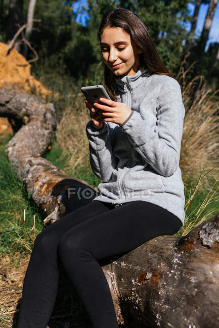 Позитивна молода жінка, яка ходить в активному одязі, сидить на стовбурі дерева і перевіряє маршрут на мобільному телефоні, проводячи сонячний день у зеленому лісі — стокове фото