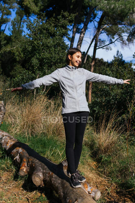 Full body of happy young female in sportswear balancing on fallen tree trunk while hiking through green forest in sunny day — Stock Photo