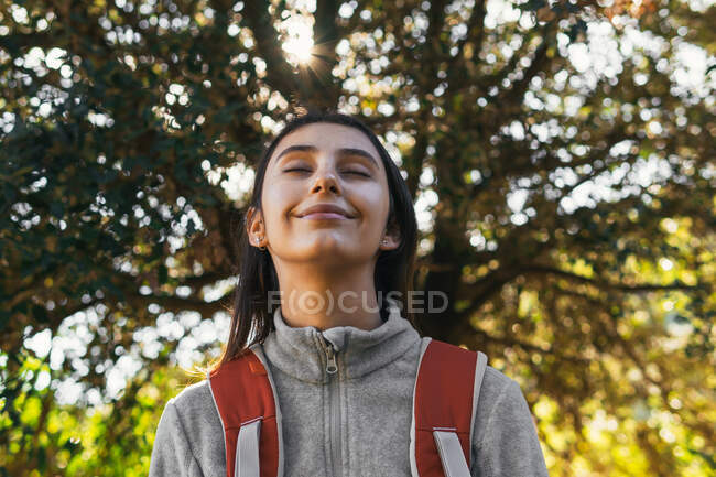 Счастливая оптимистичная молодая туристка в спортивной одежде с рюкзаком, наслаждающаяся путешествием в зеленом лесу в солнечный день с закрытыми глазами — стоковое фото