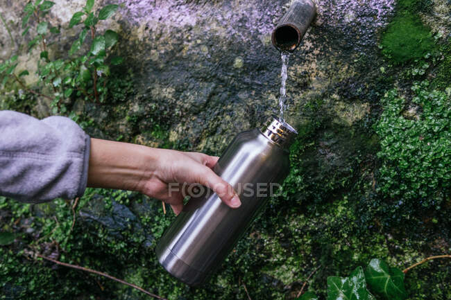 Crop anonymous hiker filling metal bottle with fresh cold natural spring water from source with pipe in mountainous forest — Stock Photo