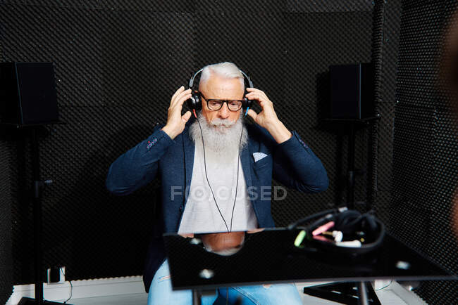 Homem idoso barbudo em fones de ouvido sentado na sala à prova de som durante o exame de audiologia e teste auditivo — Fotografia de Stock