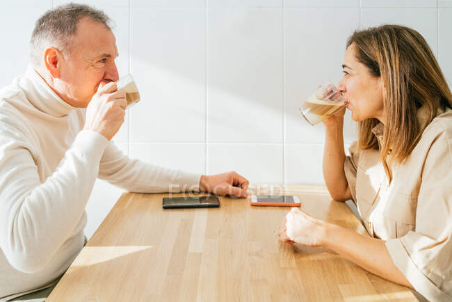 Vista lateral del contenido pareja de adultos sentados en la mesa en la cocina y disfrutando del café aromático mientras desayunan y se miran entre sí - foto de stock