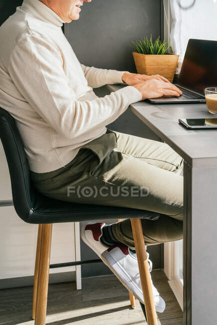 Vista lateral de corte irreconhecível macho de meia-idade trabalhando no balcão com netbook e xícara de café na cozinha pela manhã — Fotografia de Stock