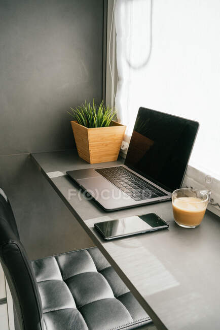 Moderno portátil y teléfono inteligente colocado en el mostrador con taza de café de la mañana en la cocina iluminada por la luz del sol - foto de stock