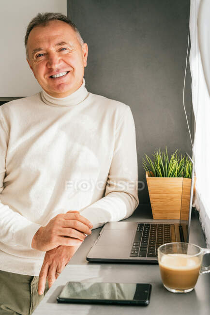 Sorrindo de meia-idade masculino inclinado no balcão com netbook e xícara de café, enquanto em pé na cozinha de manhã e olhando para a câmera — Fotografia de Stock