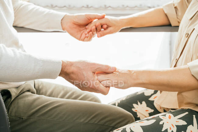 Side view of crop unrecognizable mature couple sitting at counter in kitchen and holding hands while spending time at home — Stock Photo