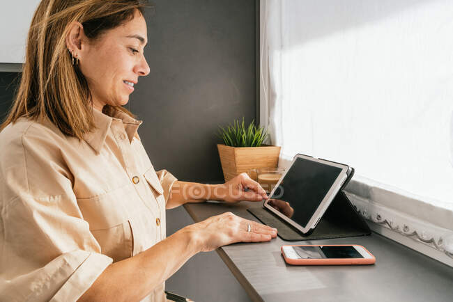 Side view of mature female sitting at counter in kitchen and browsing tablet in morning — Stock Photo