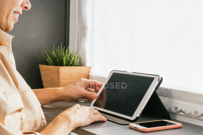 Side view of cropped unrecognizable mature female sitting at counter in kitchen and browsing tablet in morning — Stock Photo
