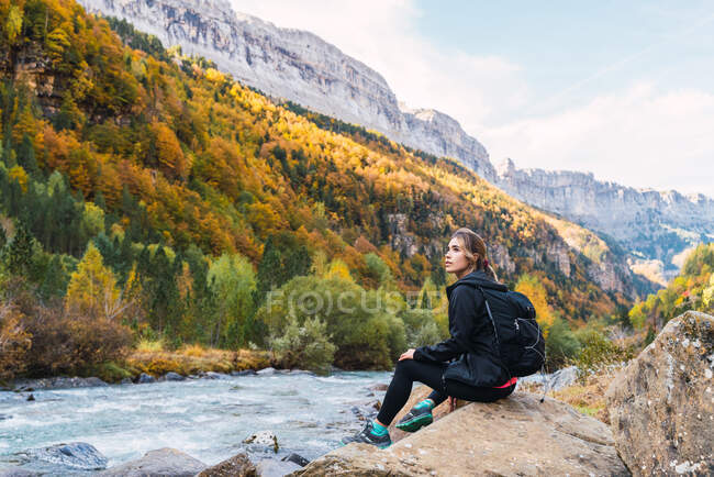 Vista lateral do caminhante feminino com mochila sentada em pedra perto do rio nas montanhas dos Pirenéus enquanto desfruta de férias no Parque Nacional Ordesa y Monte Perdido — Fotografia de Stock