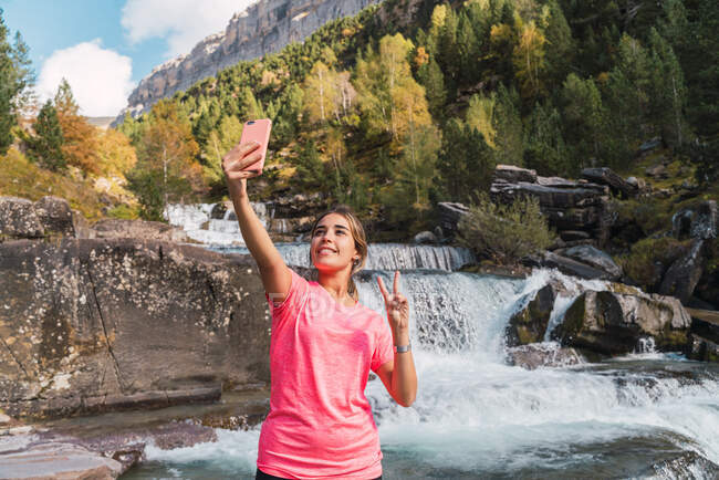 Очарованная женщина-исследователь делает самоснимок на смартфоне и показывает жест двумя пальцами, стоя на фоне быстрой высокогорной реки в Национальном парке Ордеса-и-Монте-Пердидо — стоковое фото