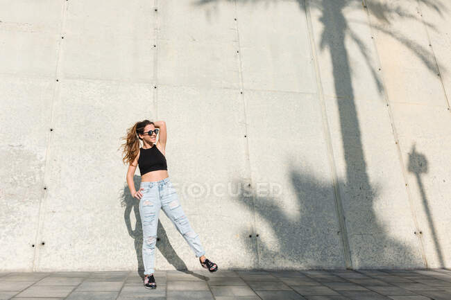 Bajo ángulo de mujer joven con estilo en gafas de sol de moda con top negro y jeans de pie cerca de un edificio moderno con sombra de palma tropical en la pared en un día soleado de verano - foto de stock