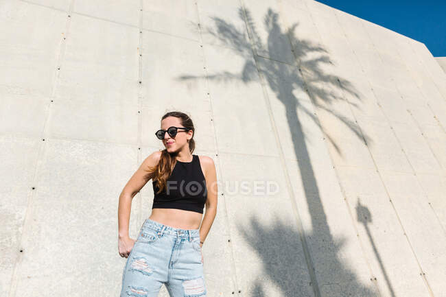 Low angle of stylish young female in trendy sunglasses wearing black crop top and jeans standing near modern building with shadow of tropical palm on wall in sunny summer day — Stock Photo