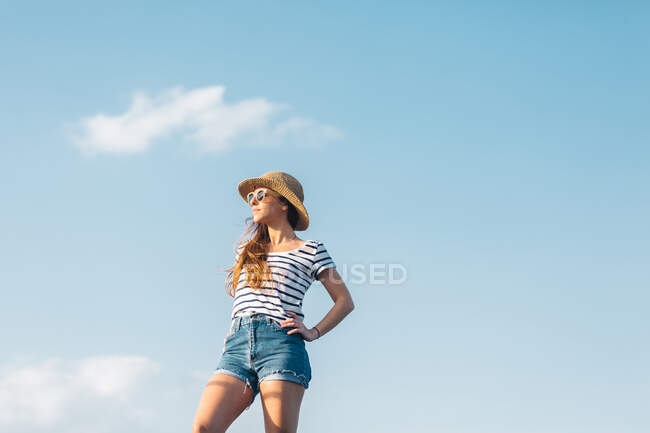 Bajo ángulo de alegre mujer joven sin preocupaciones en gafas de sol con camiseta de rayas casuales y vaqueros disfrutando de un día soleado de verano - foto de stock