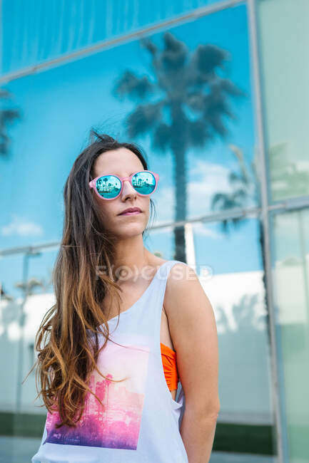 Confiant jeune femme aux cheveux longs dans des lunettes de soleil à la mode détournant les yeux tout en passant une journée ensoleillée d'été dans le parc de la ville — Photo de stock