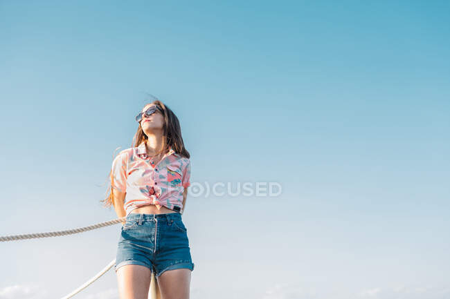 From below of modern millennial female in sunglasses wearing denim shorts and stylish casual shirt looking away while standing against blue sky in summer day — Stock Photo