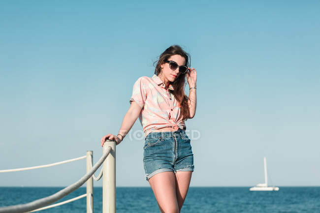 Side view of modern millennial female in sunglasses wearing denim shorts and stylish casual shirt looking away while standing against blue sky in summer day — Stock Photo