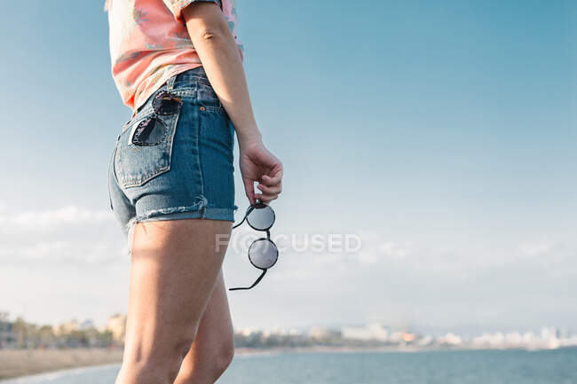 Side view of crop anonymous female teenager in denim shorts with stylish sunglasses in hand standing near sea on urban beach in sunny summer day — Stock Photo