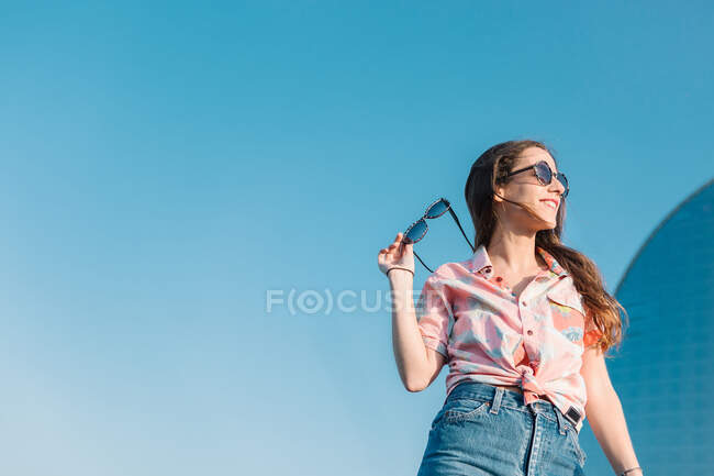 Desde abajo de la mujer milenaria moderna en gafas de sol con pantalones cortos de mezclilla y camisa casual con estilo mirando hacia otro lado mientras está de pie contra el cielo azul en el día de verano - foto de stock