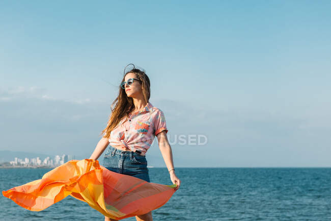 Young long haired female in trendy casual outfit and sunglasses with colorful cloth flattering on wind standing near sea water in sunny summer day — Stock Photo