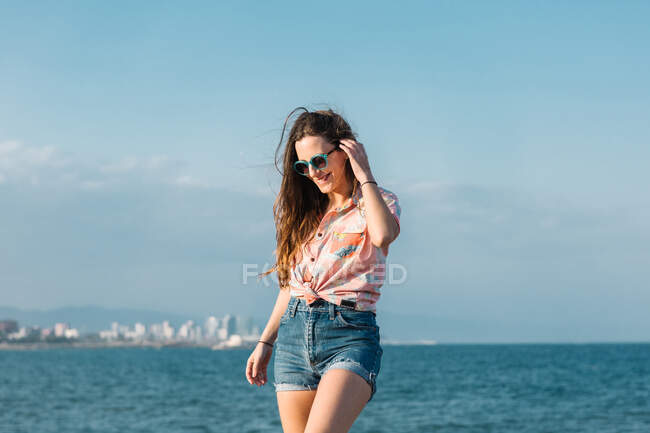 Вид на современную женщину тысячелетия в солнцезащитных очках в джинсовых шортах и стильной повседневной рубашке, смотрящую в сторону, стоя на фоне голубого неба в летний день — стоковое фото