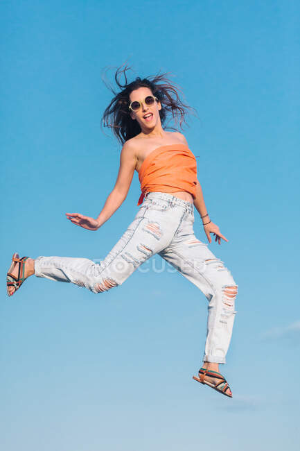 Dal basso corpo pieno di moderna hipster femminile in occhiali da sole che indossa camicia e jeans strappati alla moda che saltano in alto contro il cielo blu — Foto stock