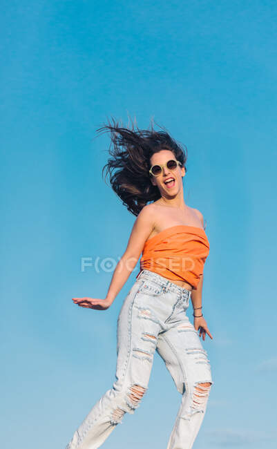 Знизу повне тіло сучасної хіпстерки-жінки в сонцезахисних окулярах в сорочці і стильні рвані джинси стрибають високо на блакитне небо — стокове фото