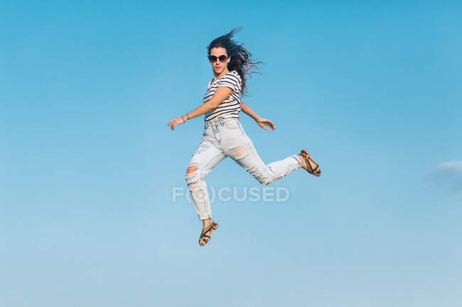 Desde abajo todo el cuerpo de la moderna mujer hipster en gafas de sol con camisa a rayas y vaqueros rasgados con estilo saltando alto contra el cielo azul - foto de stock