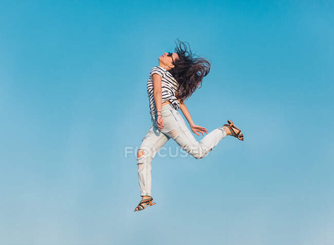 Dal basso corpo pieno di moderna hipster femminile in occhiali da sole indossando camicia a righe ed eleganti jeans strappati che saltano in alto contro il cielo blu — Foto stock
