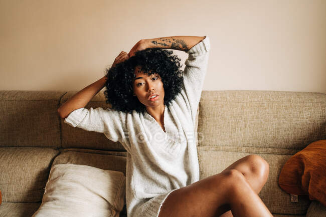 Femme afro-américaine confiante avec les cheveux bouclés assis sur le canapé et penché sur la main tout en regardant la caméra à la maison — Photo de stock