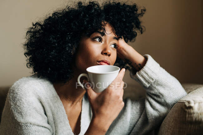 Mujer afroamericana pensativa con taza de bebida sentada en el sofá en casa y mirando hacia otro lado en contemplación - foto de stock