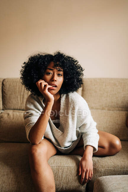 Femme afro-américaine confiante avec les cheveux bouclés assis sur le canapé et penché sur la main tout en regardant la caméra à la maison — Photo de stock