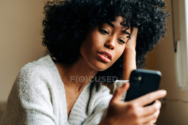Side view of a thoughtful African American female sitting on soft couch in living room and browsing mobile phone at weekend at home — стоковое фото