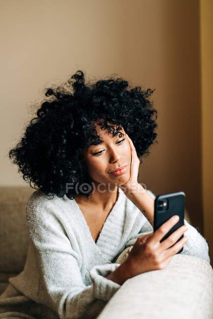 Side view of thoughtful African American female sitting on soft couch in living room and browsing mobile phone at weekend at home — Stock Photo