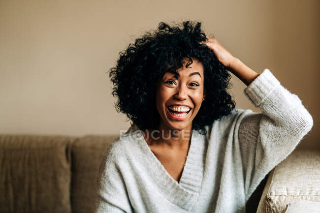 Cheerful African American female with curly hair sitting on couch touching hair while looking at camera at home — Stock Photo