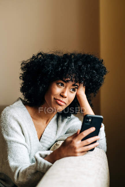 Vue latérale de la femme afro-américaine heureuse assise sur un canapé moelleux dans le salon et naviguant sur son téléphone portable le week-end à la maison — Photo de stock
