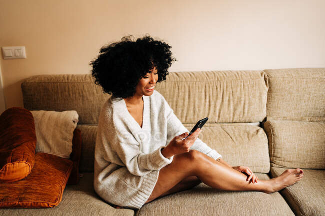 Side view of cheerful African American female sitting on soft couch in living room and browsing mobile phone at weekend at home — Stock Photo