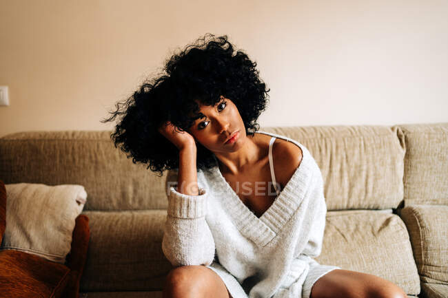 Впевнена афро-американська жінка з кучерявим волоссям сидить на дивані торкаючись волосся, дивлячись на камеру вдома. — стокове фото
