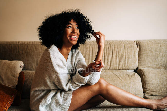 Side view of cheerful African American female with curly hair sitting on couch touching hair while looking at camera at home — Stock Photo
