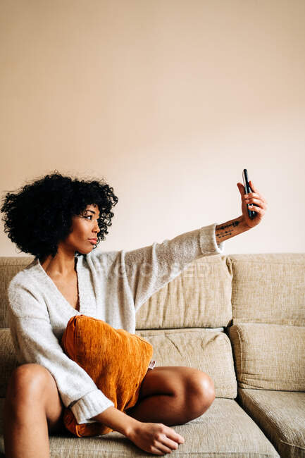 Vista lateral da fêmea étnica com penteado afro tirando selfie na câmera do smartphone enquanto desfruta de fim de semana sentado no sofá em casa — Fotografia de Stock