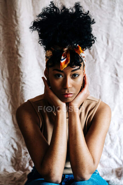 Calma femmina nera in fascia alla moda e con acconciatura Afro appoggiata alle mani e guardando la fotocamera su sfondo beige — Foto stock