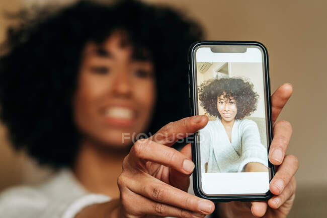 Sonriente mujer étnica con peinado afro tomando selfie en la cámara del teléfono inteligente mientras disfruta de fin de semana en casa - foto de stock