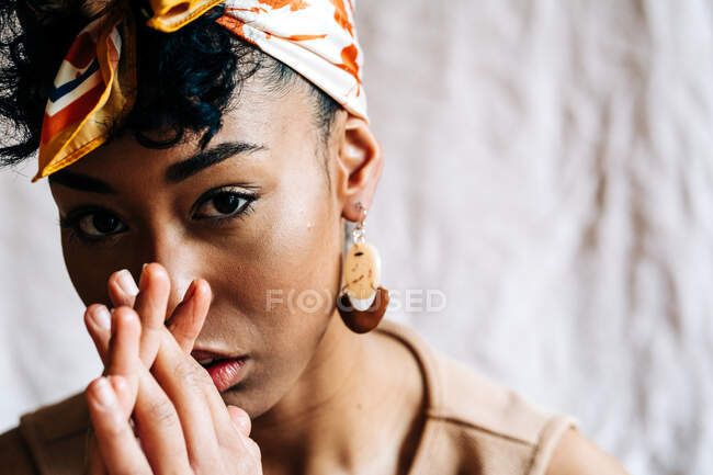Calm black female in trendy headband and with Afro hairstyle leaning on hands and looking at camera on beige background — Stock Photo