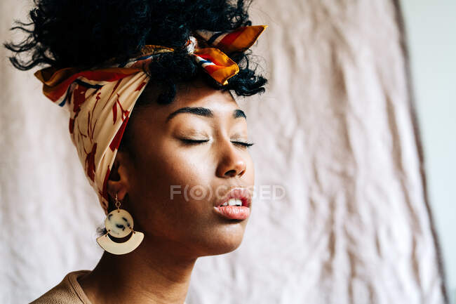 Mulher afro-americana despreocupada na cabeça elegante e com penteado afro contra tecido bege em casa — Fotografia de Stock