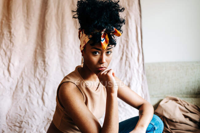 Vista lateral calma mulher preta na moda headband e com penteado afro apoiando-se nas mãos e olhando para a câmera no fundo bege — Fotografia de Stock