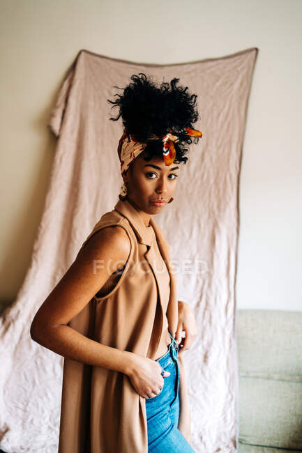 Vista lateral de la mujer afroamericana en diadema y ropa de moda de pie en la habitación y mirando a la cámara - foto de stock